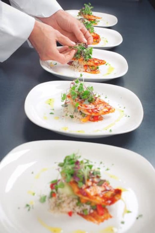 "A Chef's hands working the line plating and putting the finishing touches on chicken entrees. Focus is on the hands. Entree is Grilled BBQ Chicken served with spicy brown rice, steamed vegetables and microgreens."